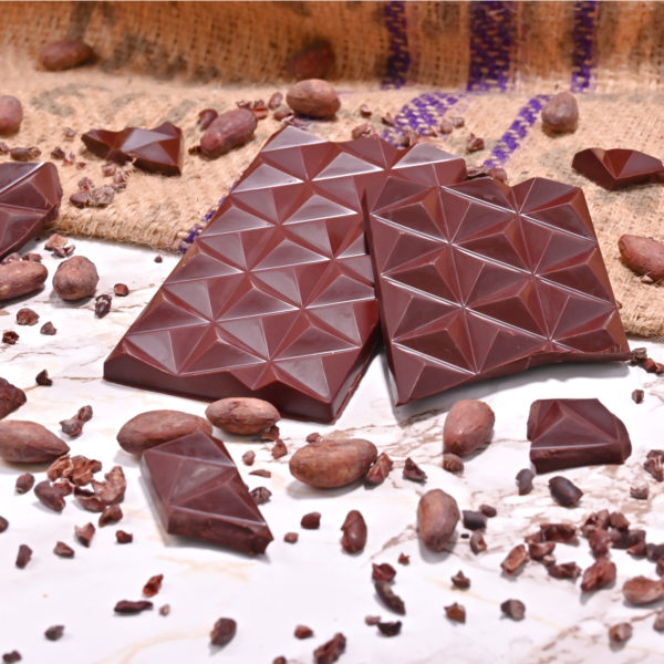Foto einer dunklen Schokoladentafel von Chani Schoggi in Luzern. Die zwei Tafeln liegen auf einem Jutesack und Marmorboden und sind umgeben von Kakaonibs und Kakaobohnen. Die Schokolade wurde Bean-to-Bar hergestellt.